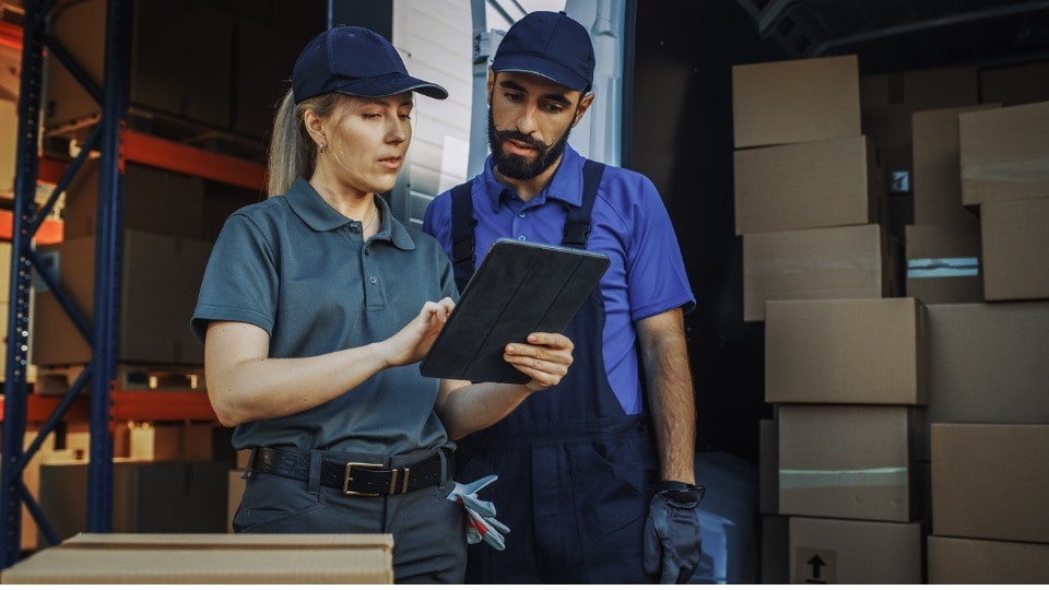 outside of a logistics distributions warehouse with manager using a tablet, talking to employee loading delivery truck with packages