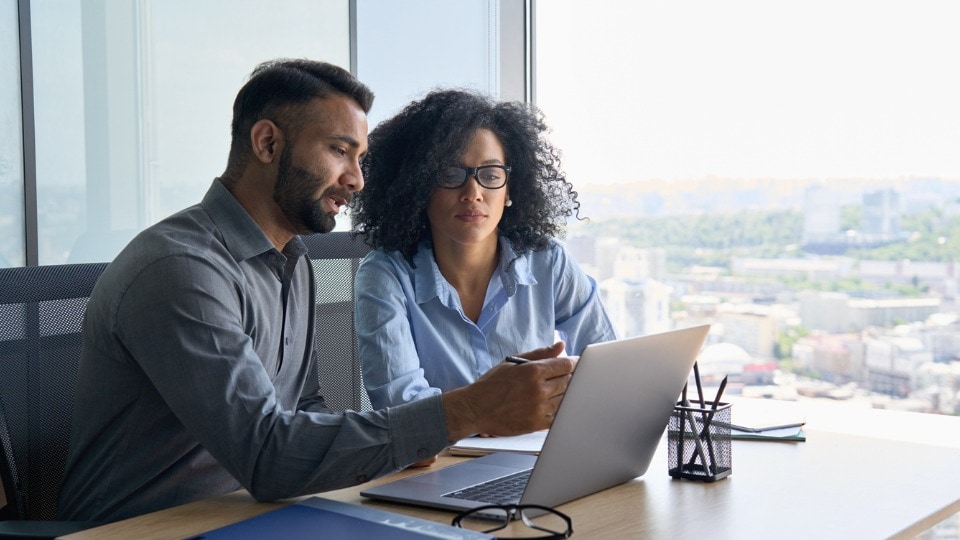 Two colleagues sitting at desk looking at laptop computer in office